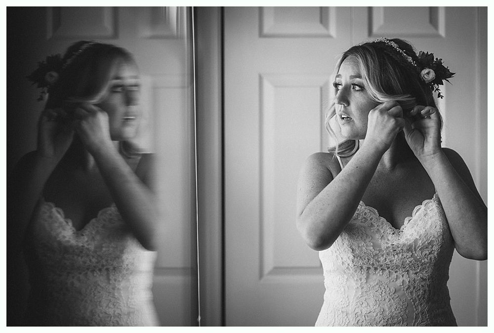 Bride adjusting earring, wearing a lace bodice dress, standing near a mirror.