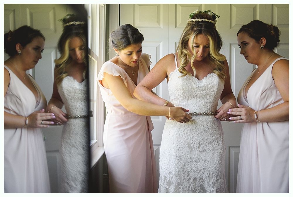 Bride having her belt adjusted by bridesmaid. Other bridesmaids observe in a room.