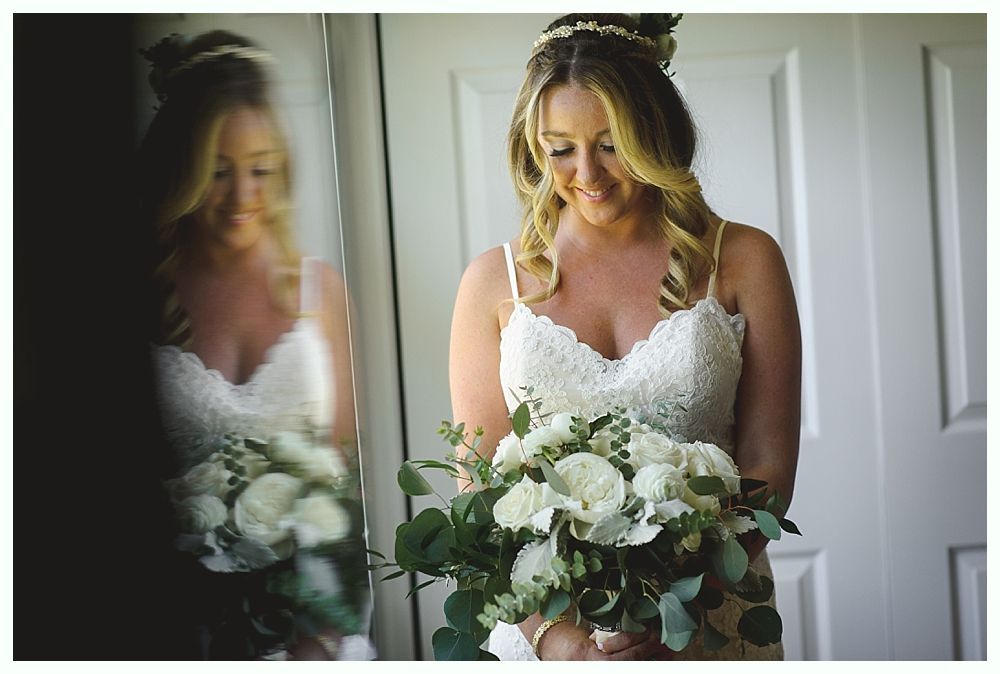 Bride in white lace dress holding bouquet, smiling by a window, reflection in mirror.