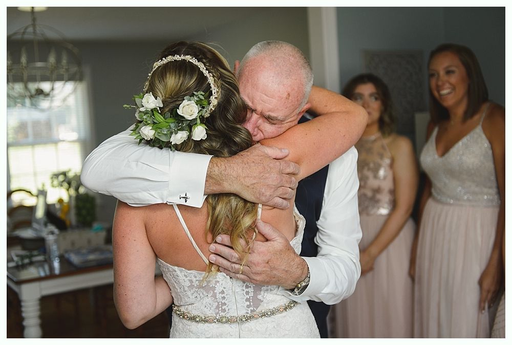 Bride in wedding dress embraces older man, with bridesmaids watching.
