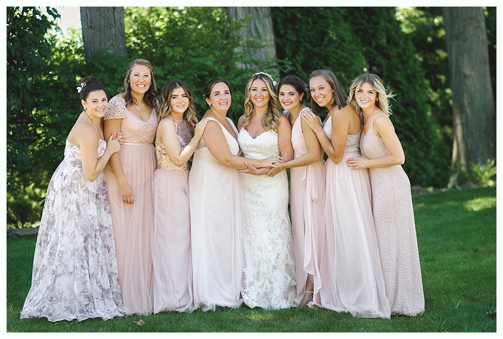Bridesmaids in pink dresses surround a bride in a white wedding gown on a lawn.