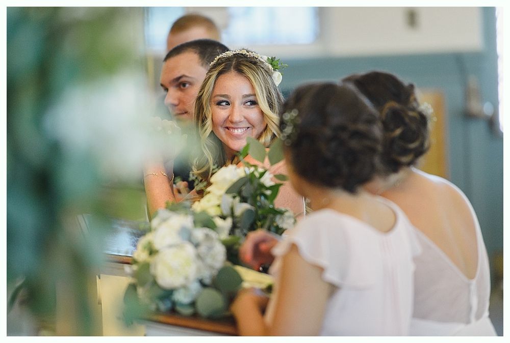 Bride smiles, holding a bouquet, with bridesmaids and groom in a church.