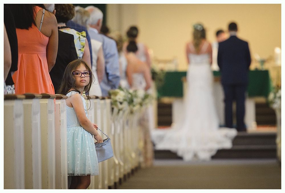 Girl with glasses holding a basket stands in aisle at a wedding. Bride and groom in background.