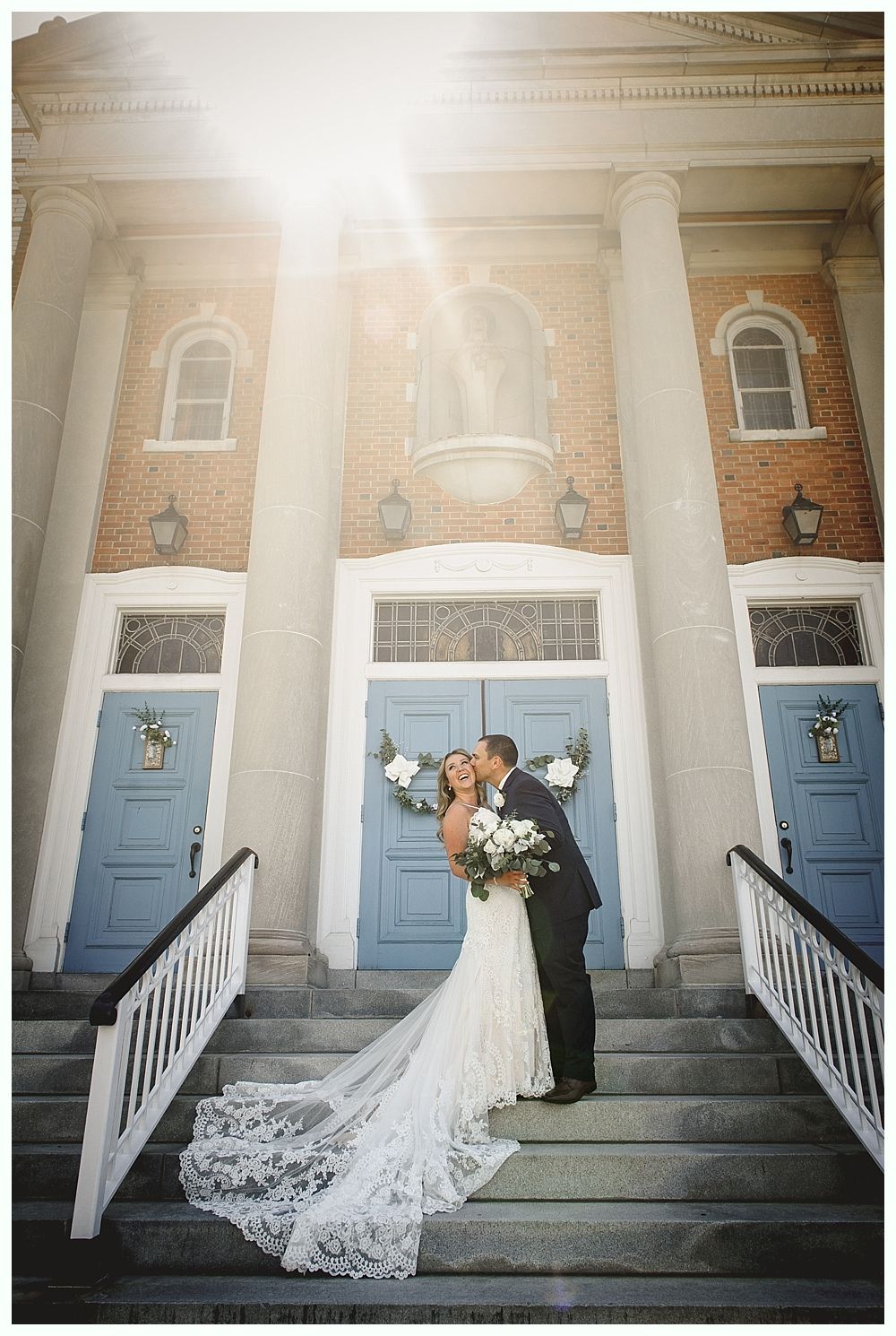 Couple kisses on steps in front of a church with blue doors, sunlight beaming down. Bride in white dress, groom in suit.
