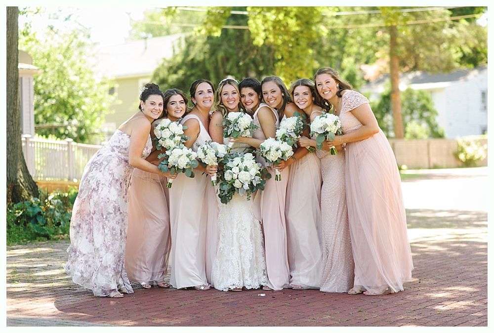 Bridesmaids in blush dresses with bride holding bouquets. Outdoor setting with trees and brick path.