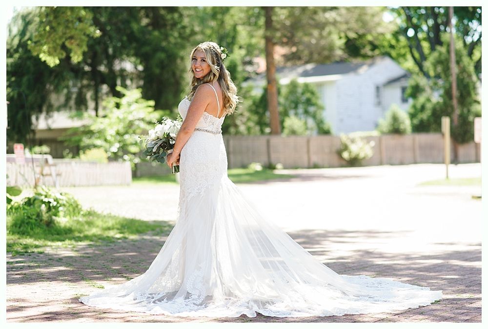Bride in white lace wedding dress with long train, holding bouquet, smiling outdoors.
