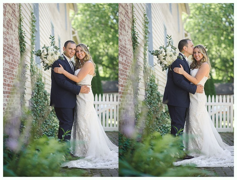 Bride and groom embracing, posing near white picket fence and building; man kisses woman's cheek.