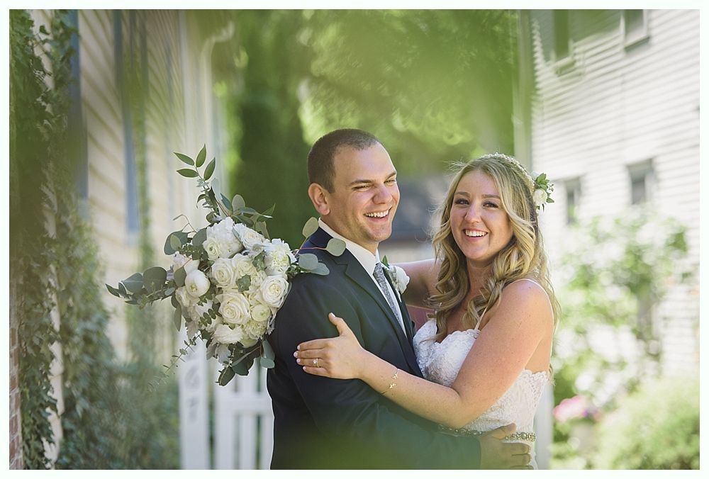 Bride and groom embrace and laugh outdoors. Bride wears white dress, groom in suit, holding a bouquet.