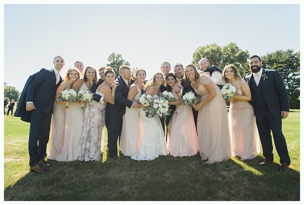 Wedding party posing on a green lawn, sunny day. Bridesmaids in blush gowns, groomsmen in navy suits.