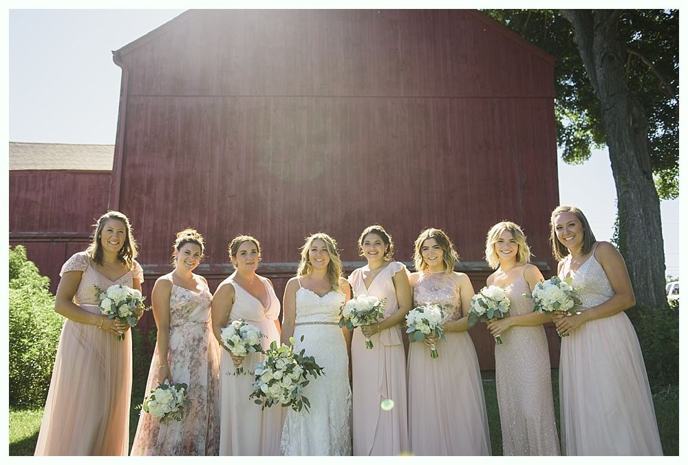 Bridesmaids and bride pose in front of a red barn, holding bouquets. They wear pink and white dresses.
