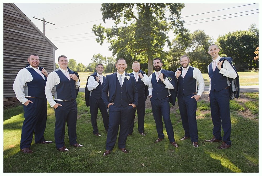 Groomsmen in navy suits pose outdoors near a barn, smiling.