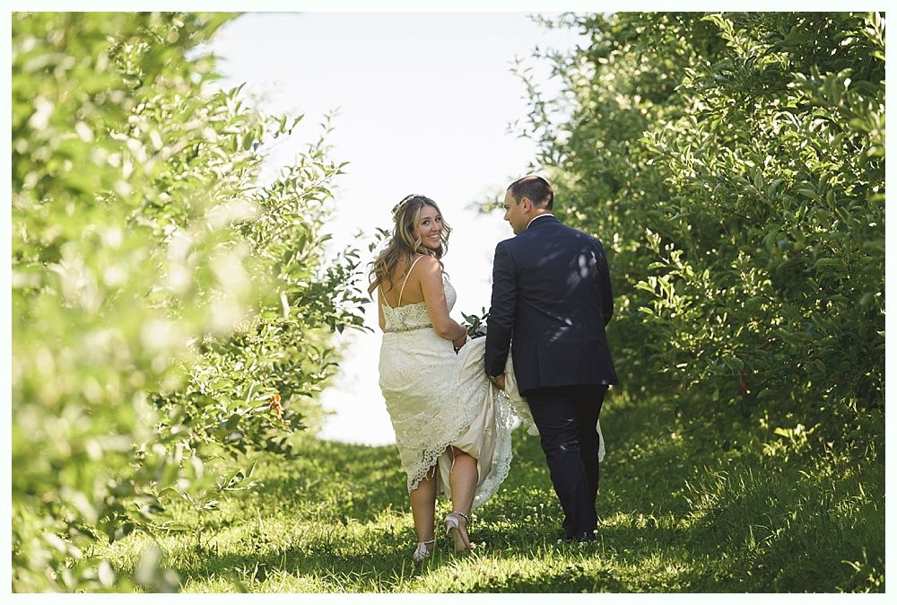 Bride and groom walk hand in hand through sunlit orchard, smiling, facing away.