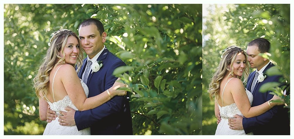 Newlyweds embrace outdoors, posing for a photo. The bride smiles, the groom kisses her cheek. Green foliage surrounds them.