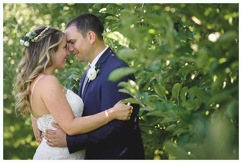Couple embraces outdoors, bride in white dress, groom in blue suit, green foliage in background.