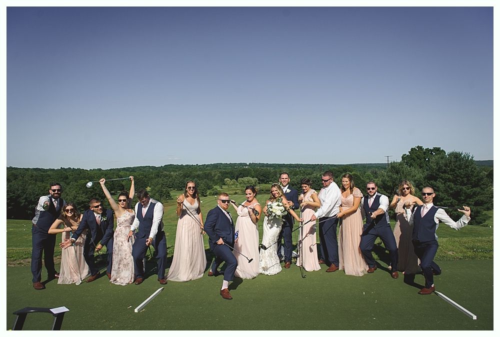 Wedding party on a golf course, posing with golf clubs. Some raise hands, wearing formal attire, sunny day.