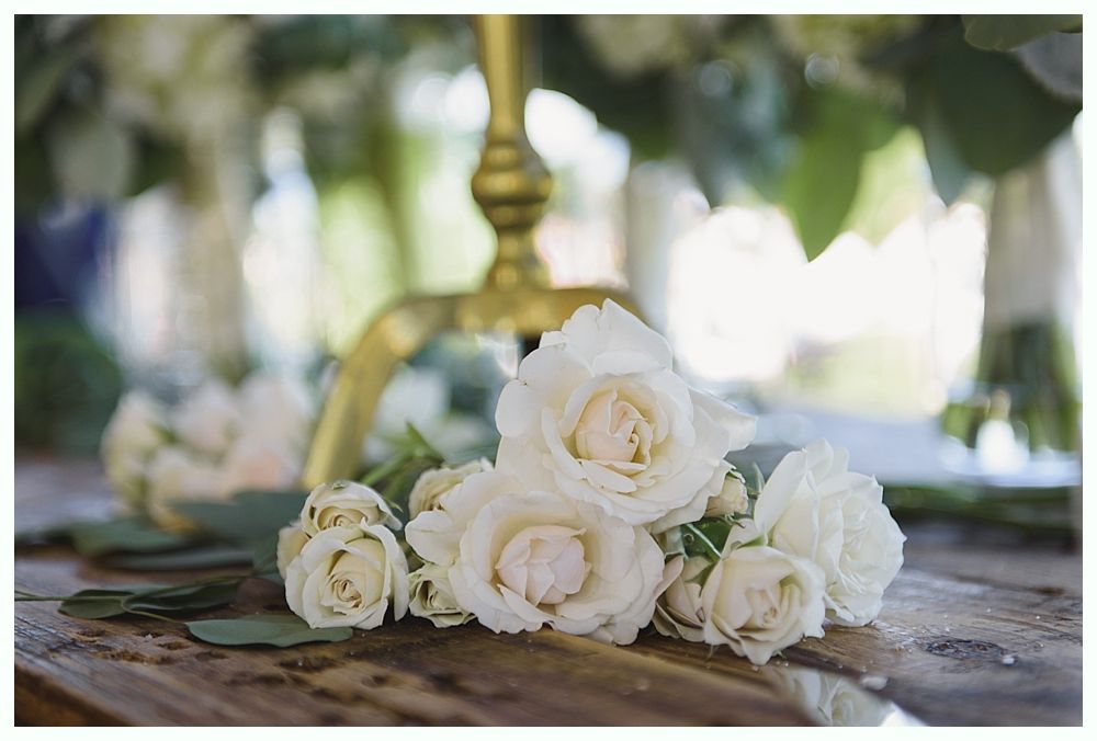 White roses on a wooden table, with a blurred gold decorative stand and greenery in the background.