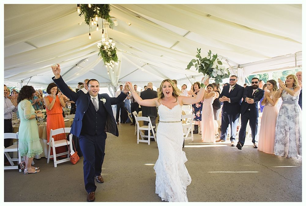 Newlyweds cheer, arms raised, exiting wedding reception tent, followed by clapping guests.