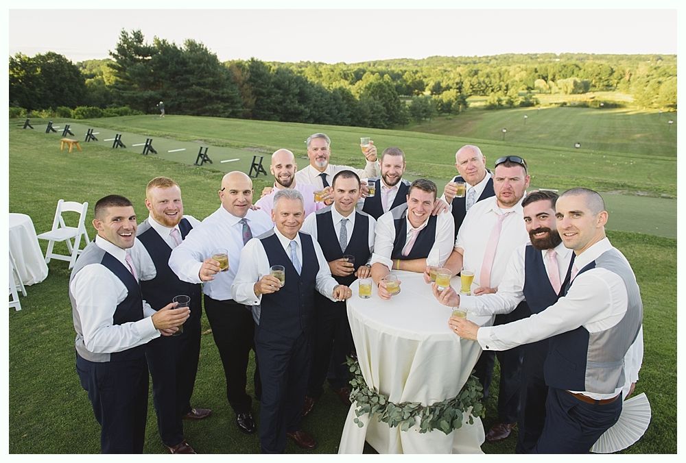 Groomsmen in vests and dress shirts toast around a small table on a grassy hill.