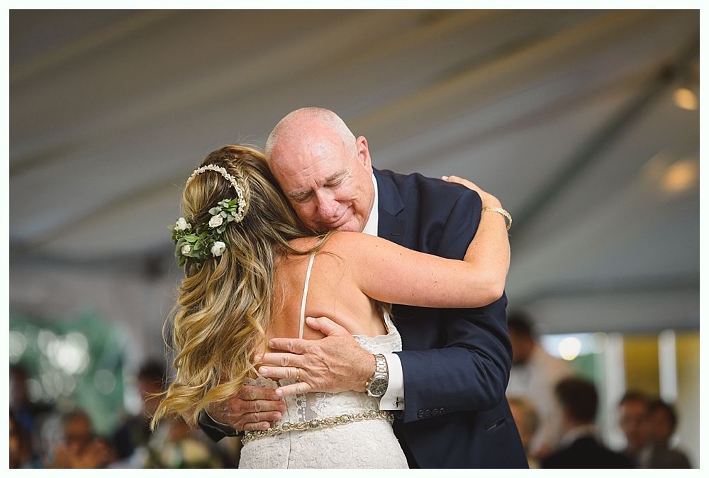 Father and bride embrace on a dance floor, he wearing a suit, she in a white dress with flower crown.