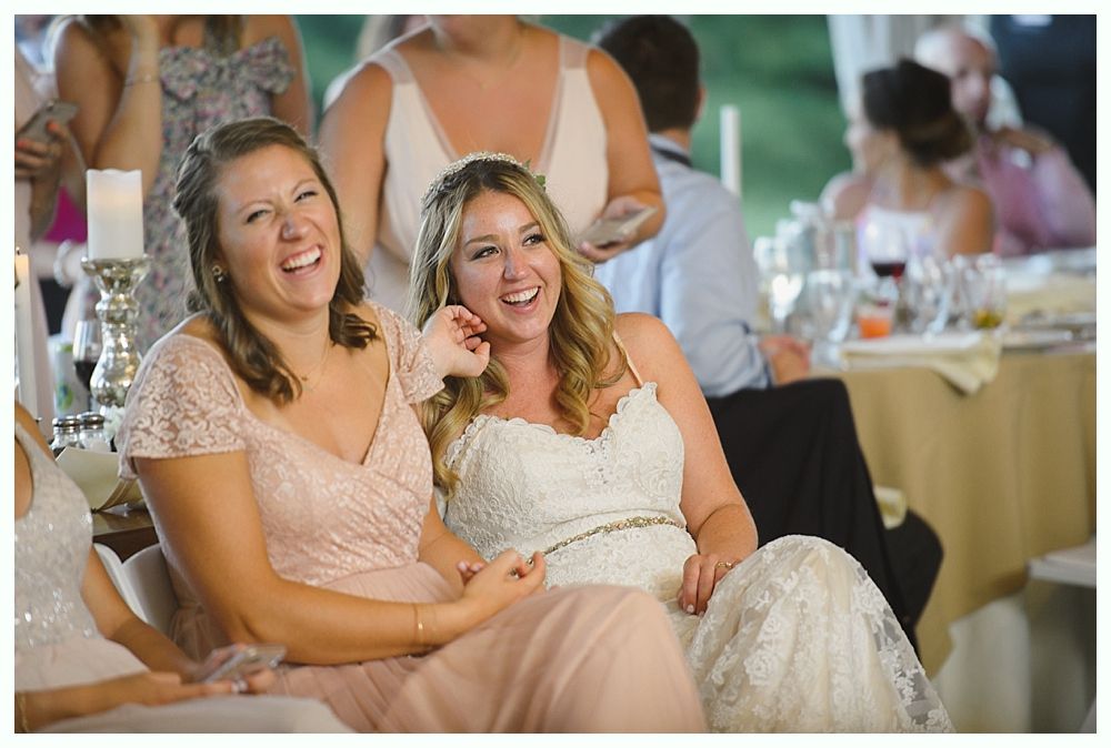 Bride and bridesmaid laughing at a wedding reception. Both are wearing formal dresses, sitting at a table.