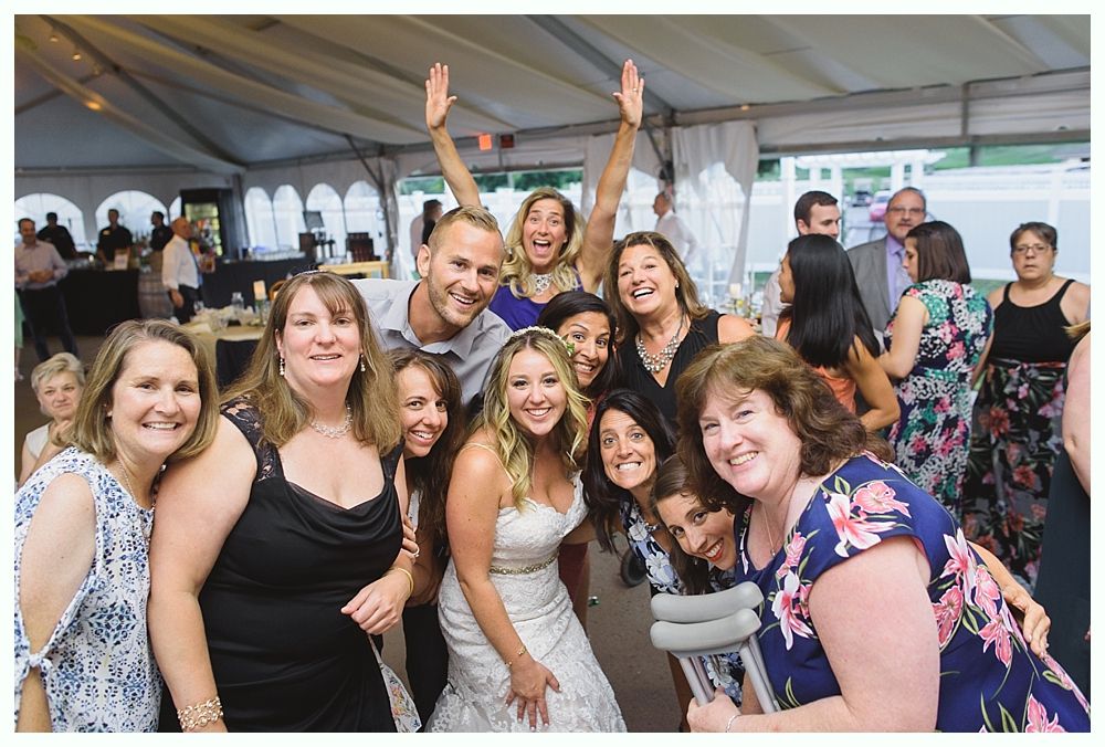 Group of people celebrating, bride in center with arms around her. Tent setting. Joyful expressions.