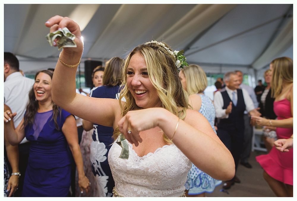 Bride dancing, holding money, smiling joyfully at a wedding reception tent. Other guests are dancing.
