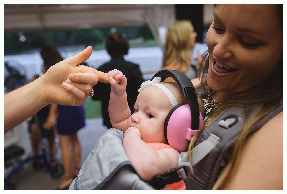 Baby wearing earmuffs and held by smiling woman; outdoors, finger touching baby's hand.