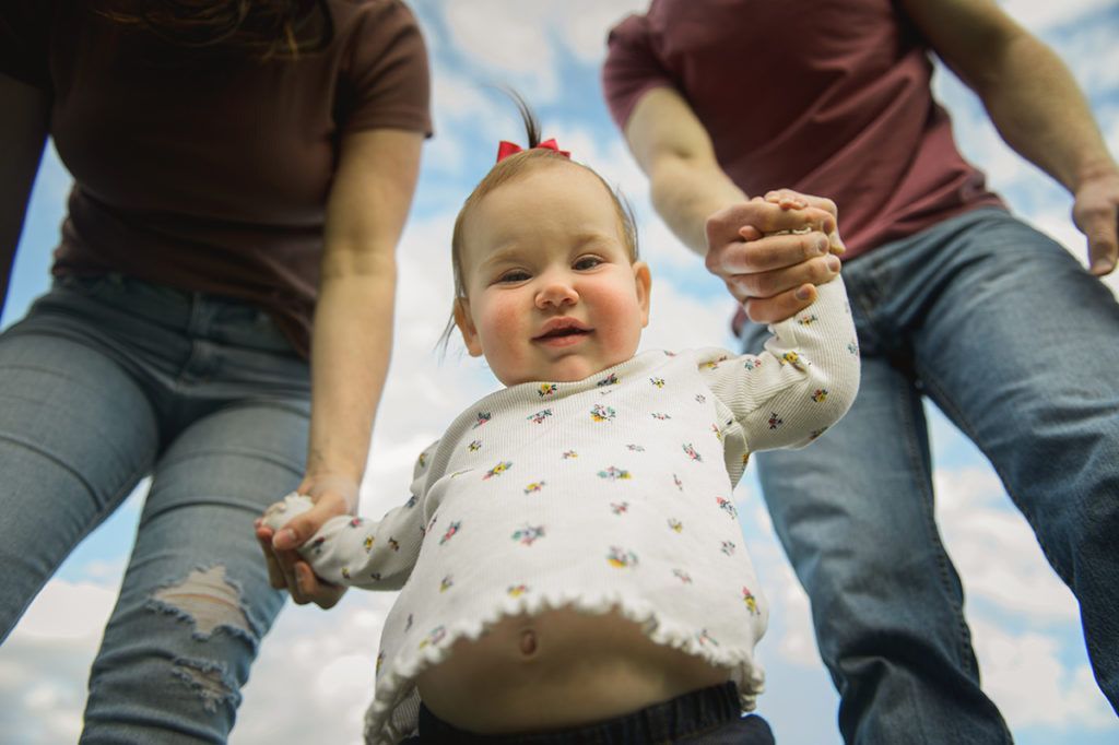 Child learning to walk, held by parents' hands, looking up with a smile. Blue sky background.