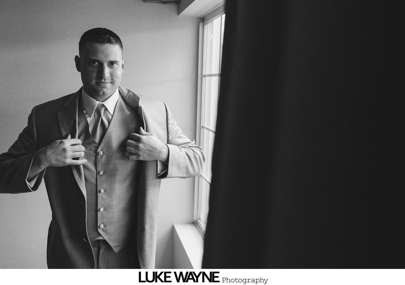 Man in a suit adjusting his jacket, standing near a window. Black and white.
