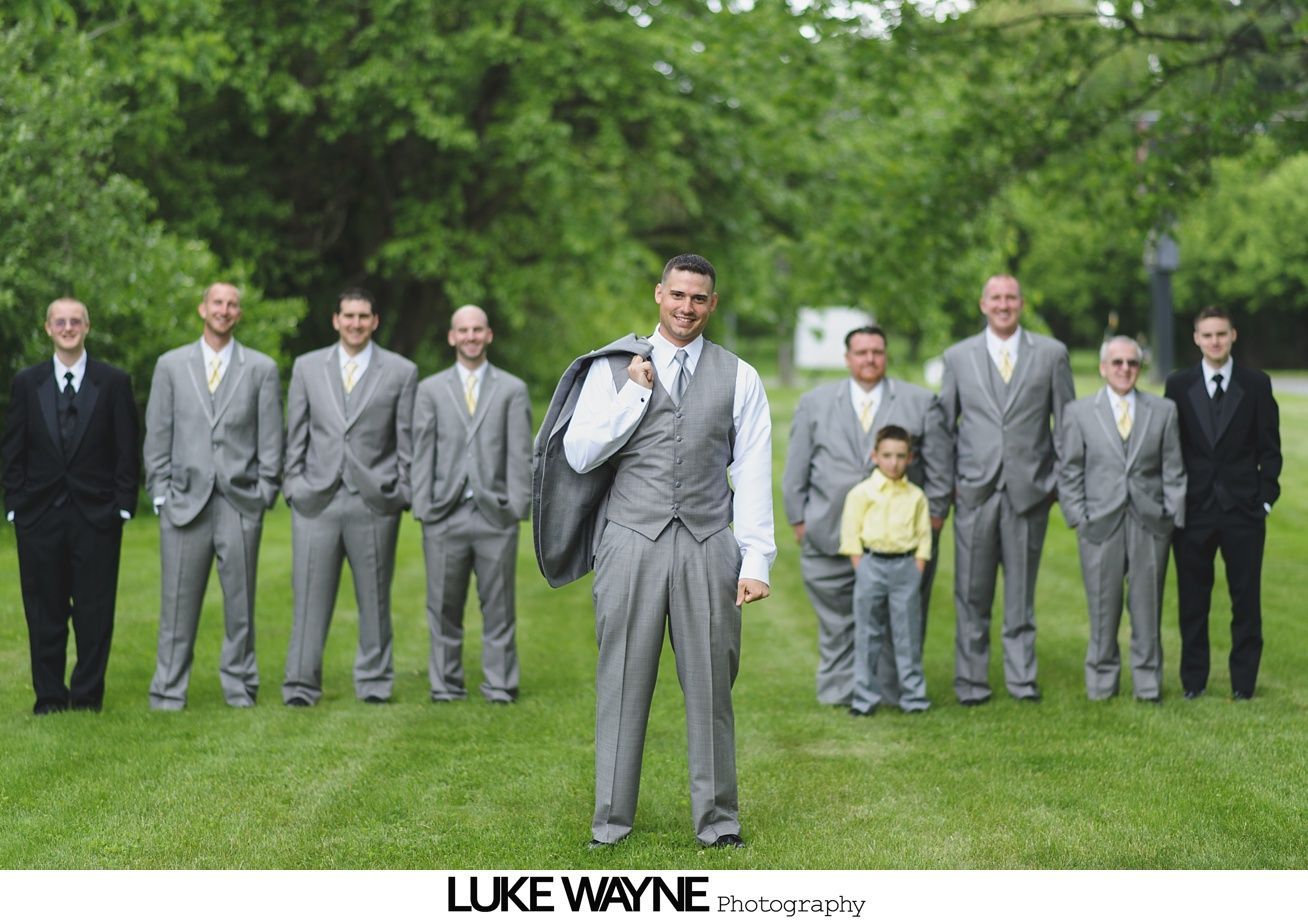 Men in grey suits pose on green grass, groom holding coat.