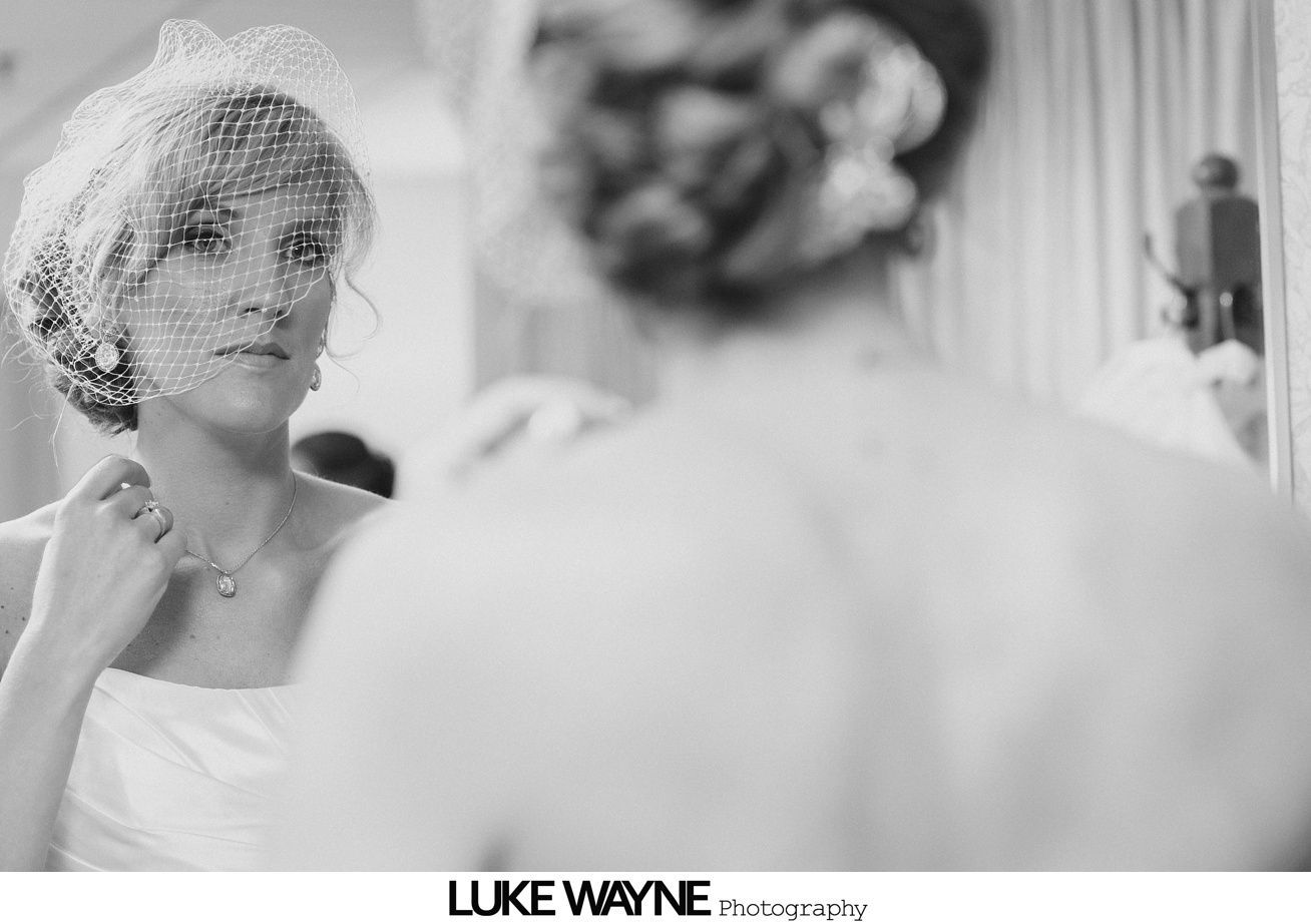 Bride in wedding dress, looking in mirror, adjusting necklace, soft focus on her back.