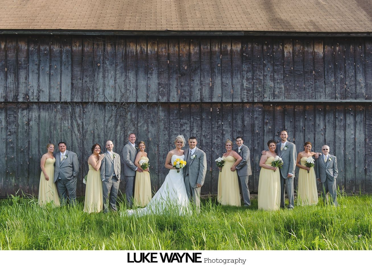 Wedding party in front of a rustic barn. Bride in white dress, bridesmaids in yellow, groomsmen in gray.