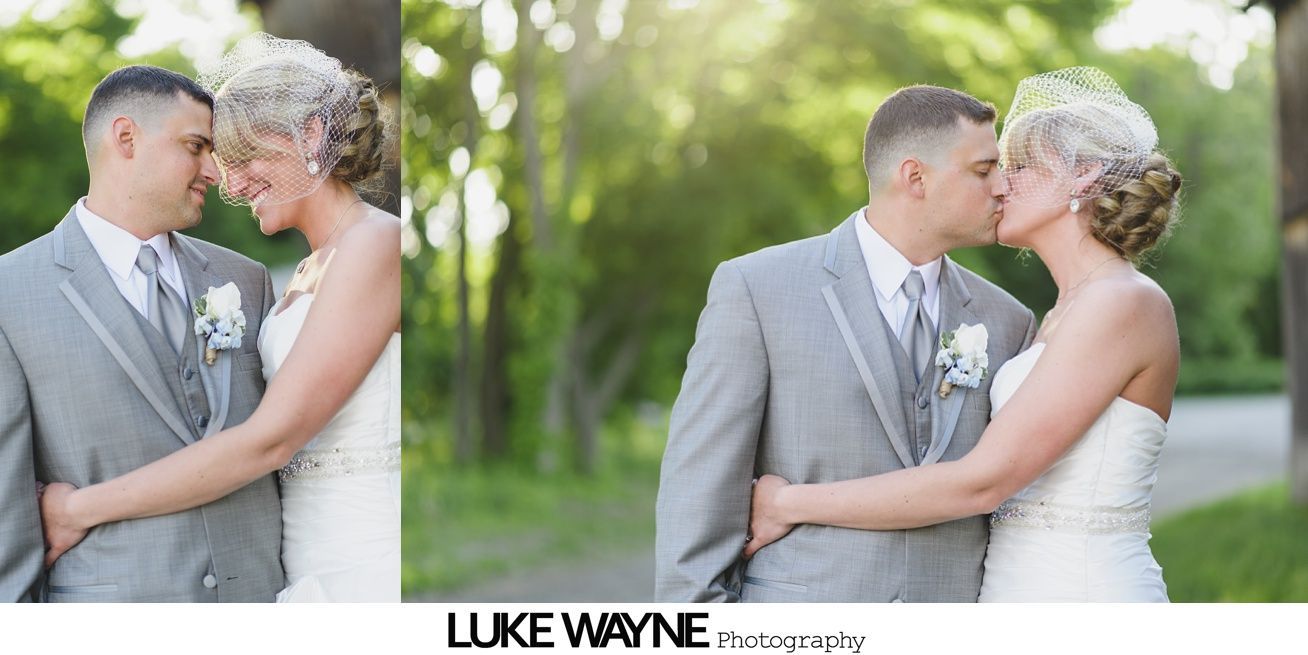 A couple embraces and kisses outdoors. The man wears a gray suit, and the woman a white dress.