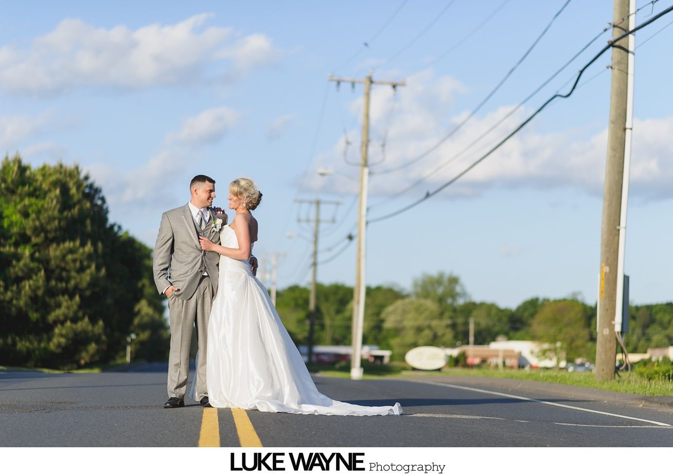 Couple in wedding attire embrace on a road with power lines in the background. Sunny day.