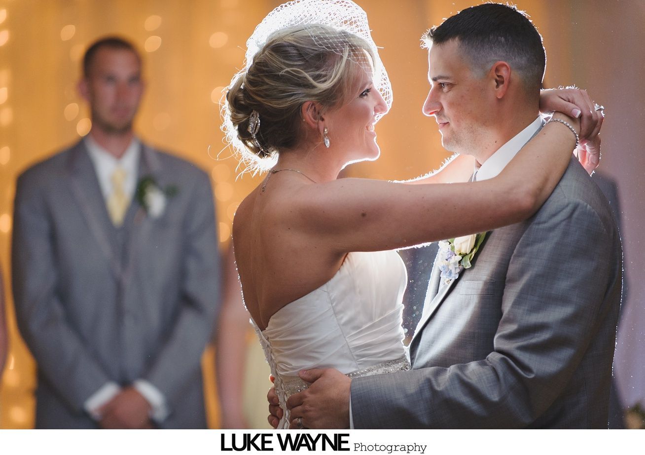 Bride and groom embrace, dancing. Man in grey suit watches from background.