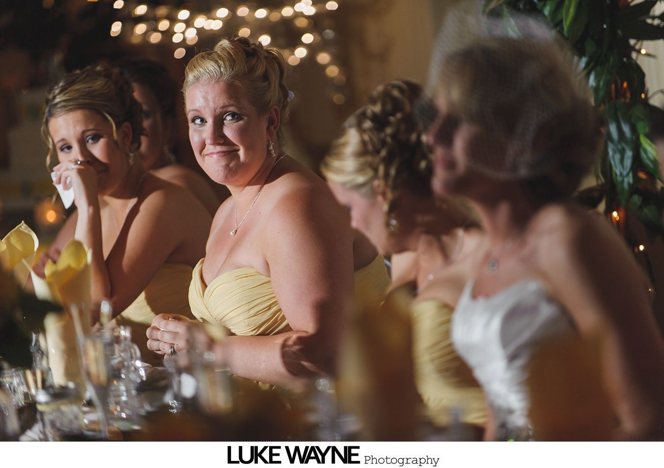Bridesmaids in yellow dresses at a wedding. One cries, others smile, and all face toward the camera, indoor setting.