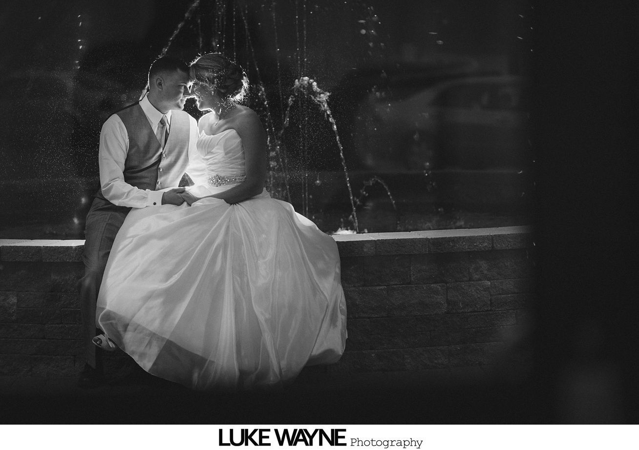 Bride and groom, illuminated by a fountain, sit close and touch faces in a romantic embrace.
