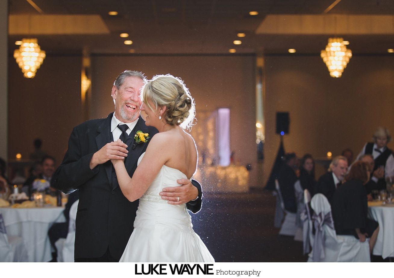 Bride and her father dance at a wedding reception. They smile, warmly lit in a ballroom with chandeliers.