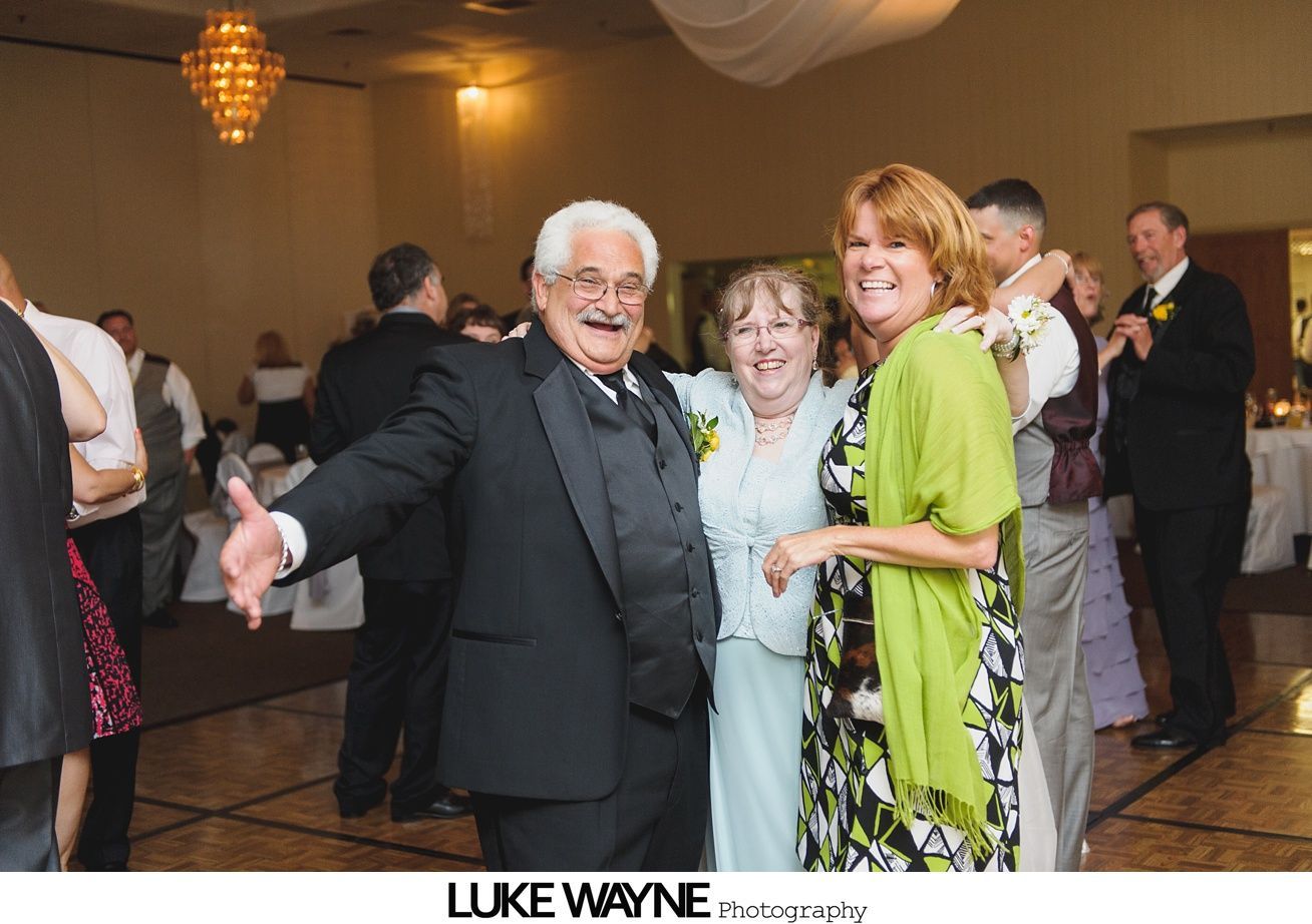 People dancing and smiling at an event, man in a tuxedo smiles, woman in a floral dress, other guests in background.