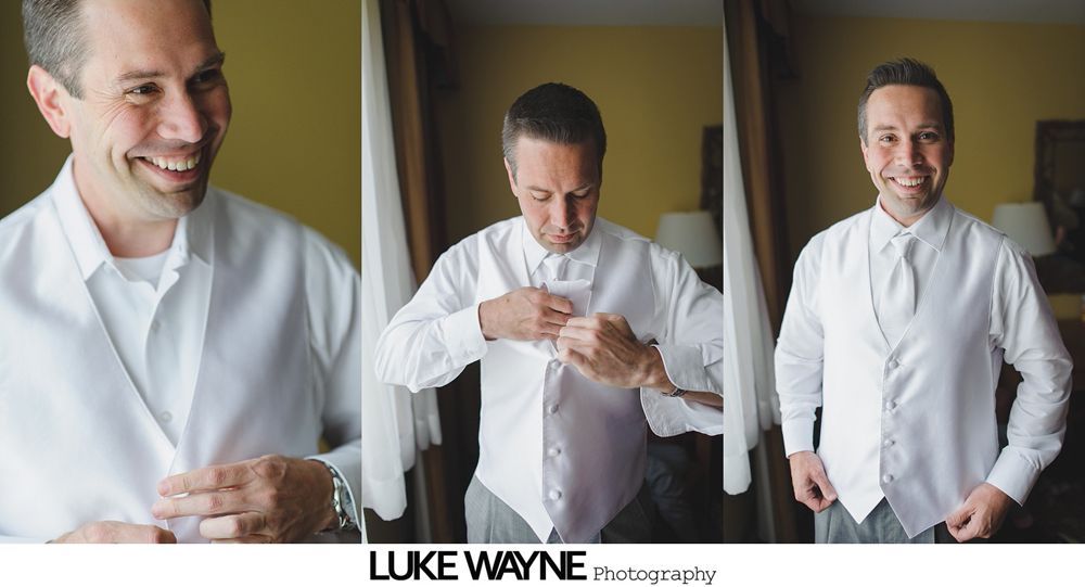 Man in a white shirt and vest putting on a button, smiling. Indoor setting with curtains.