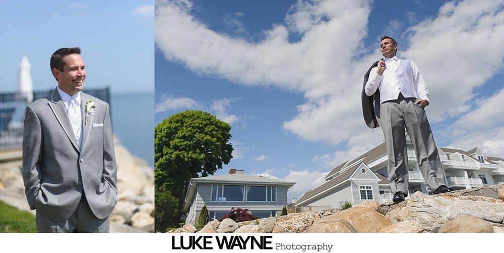 Groom in gray suit stands on a rocky hill with jacket over his shoulder, blue sky background.