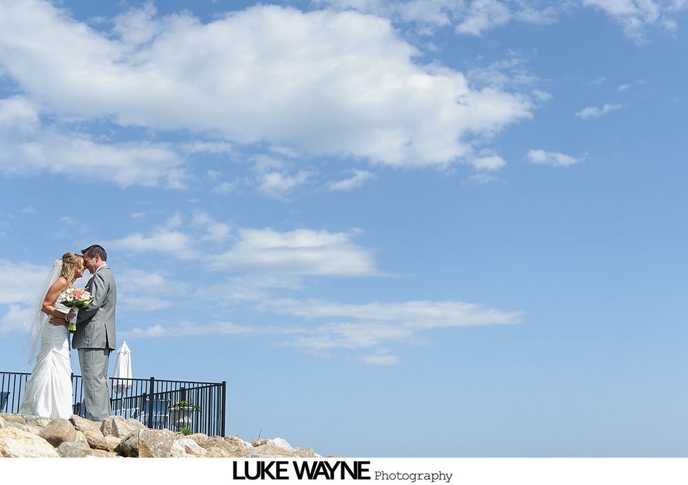 Bride and groom stand on rocks, looking at each other, against blue sky with clouds.