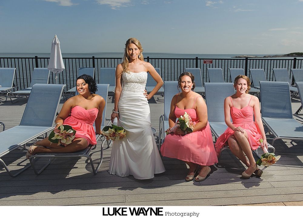 Bride with bridesmaids in coral dresses pose on lounge chairs by water.
