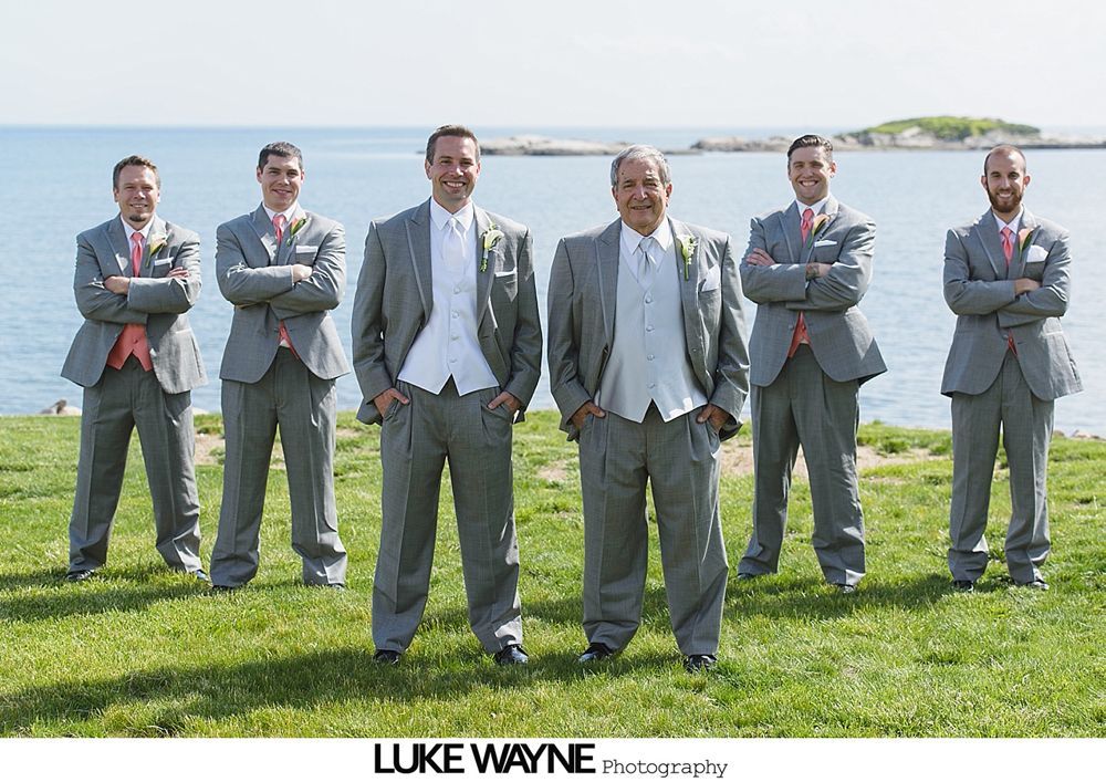 Men in gray suits, some with coral accents, pose on a grassy knoll by the ocean.