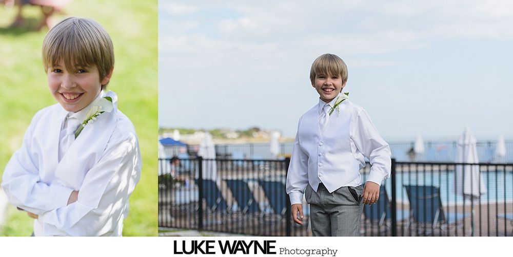 Young boy smiles while wearing a white shirt, vest, and gray pants, set against a blurred outdoor backdrop.