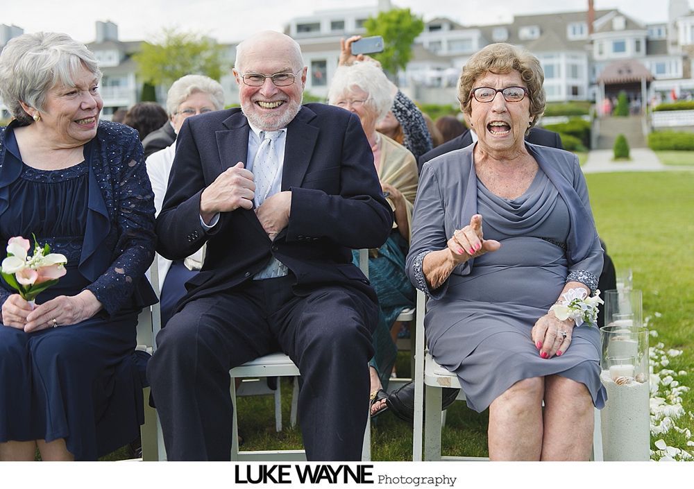 Three people seated at an outdoor event, reacting with smiles and excitement; water and buildings in the background.