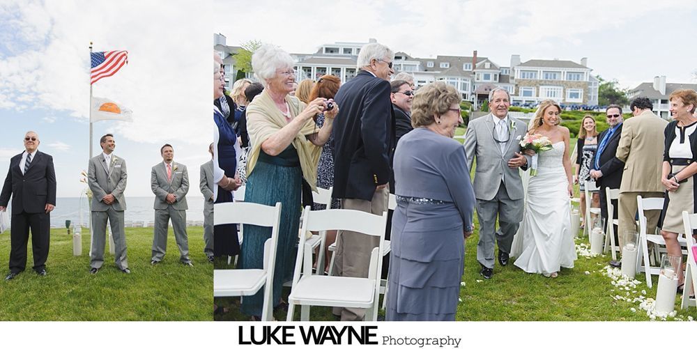 Wedding ceremony, bride escorted down the aisle. Guests watch on the grass next to the ocean, American flag in the background.