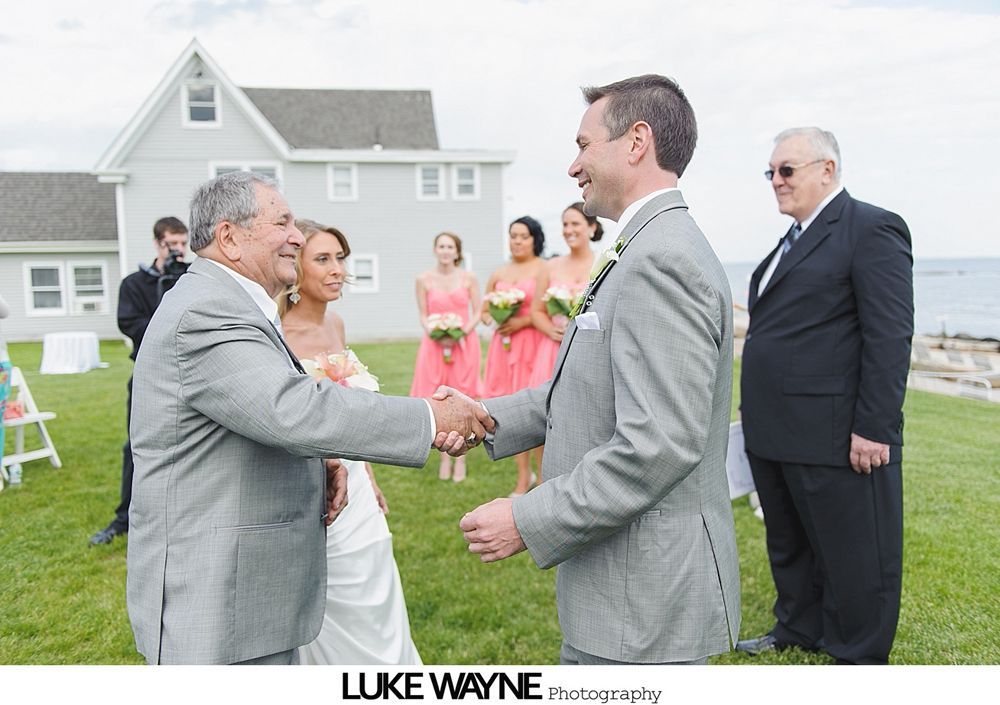Groom shakes hands with a man at an outdoor wedding. Bride, bridesmaids, and guest stand nearby.