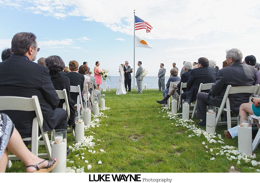 Wedding ceremony on a grassy hill with ocean view, white chairs, and American flag.