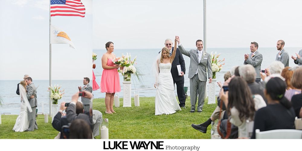 Wedding ceremony by the water, couple raises hands in celebration. Guests applaud. American flag flies.